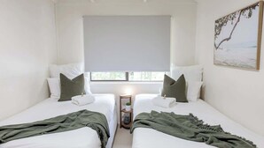 A room with two beds, a window with a view, a bedside table with a lamp, and a painting on the wall. - Cute Little Cove gem…walking distance to beach! (Noosa Heads)
