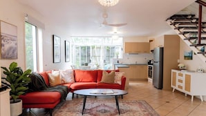 A modern living room with a red sofa, a coffee table, and a dining area with a table and chairs. There is a kitchen with cabinets and a refrigerator. - Cute Little Cove gem…walking distance to beach! (Noosa Heads)