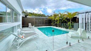Ein moderner Poolbereich im Freien mit Glasverkleidung, weißen Liegestühlen und einem Holzzaun im Hintergrund.