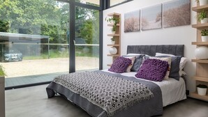 A modern bedroom with a large bed, a view of a car through glass doors, and a beach-themed wall art. - The Bridge House  -  Luxury Cottages (Bursledon)