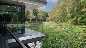 A modern outdoor deck with glass railings overlooking a body of water and lush greenery. - The Bridge House  -  Luxury Cottages (Bursledon)