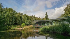 A modern building with large glass windows, situated by a calm lake surrounded by dense greenery. - The Bridge House  -  Luxury Cottages (Bursledon)