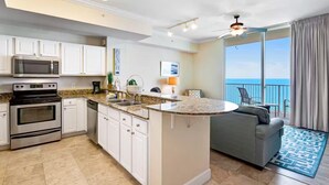 A modern kitchen with white cabinets, a built-in microwave, and a large island. There is a gray sofa and a balcony with a view of the ocean. - Tidewater 3006 (Panama City Beach)
