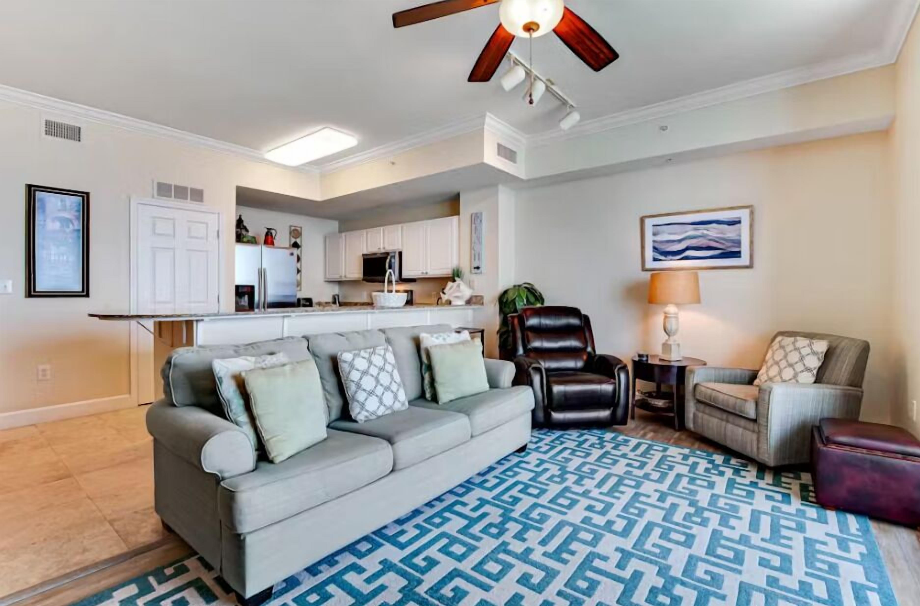 A living room with a gray sofa, a patterned rug, a ceiling fan, and a kitchen in the background.