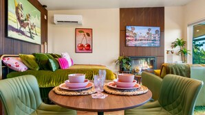 Quarto de hotel moderno com uma cama, poltronas estofadas em verde, uma mesa de madeira redonda com pratos cor-de-rosa e uma televisão na parede.