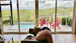 Uma sala de estar espaçosa com uma grande porta de vidro deslizante que dá para uma varanda com piscina, cadeiras vermelhas e vista para uma paisagem montanhosa.