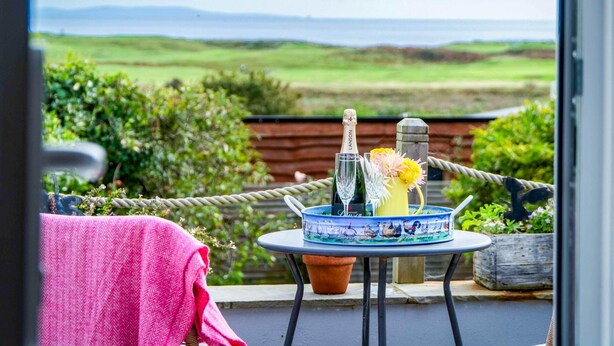 A balcony with a table set for two, featuring a bottle, glasses, and a decorative vase with flowers. - The Studio At Raymond House - Penally - Tenby (Tenby)
