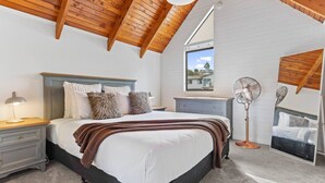 Camera da letto con soffitto in legno, un letto con biancheria da letto bianca e una coperta marrone, un comodino con una lampada, una cassettiera e una ventola da terra.
