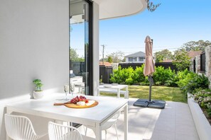 A patio with a white table, two white chairs, a small potted plant, a fruit platter, and a wine glass. - Stylish Coastal Retreat Near Cronulla Beach (Burraneer)
