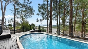 A swimming pool with a wooden deck, lounge chairs, and a view of trees and buildings.