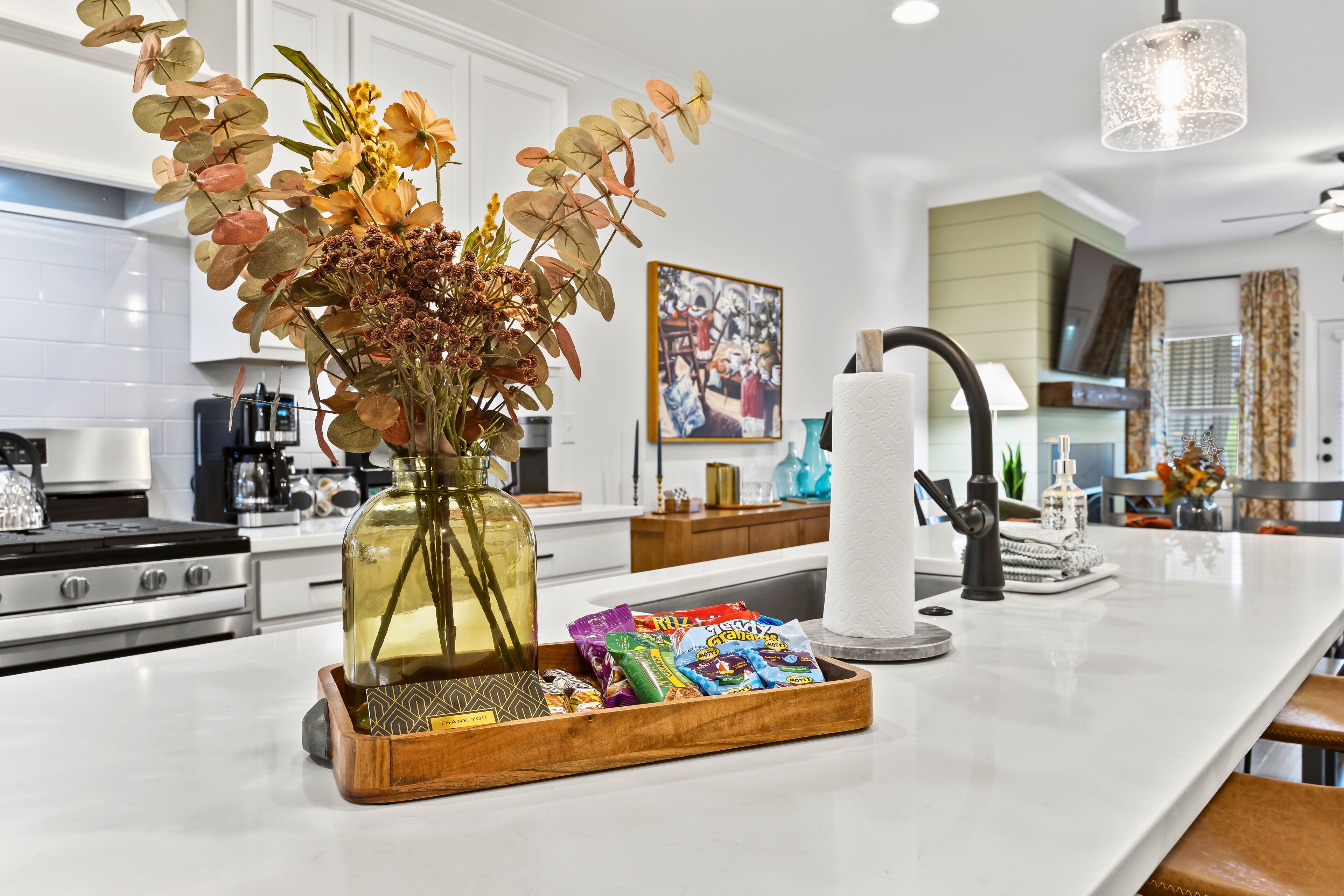 Una cocina con una isla central, un jarrón con flores secas, una bandeja con aperitivos y un dispensador de toallas de papel.