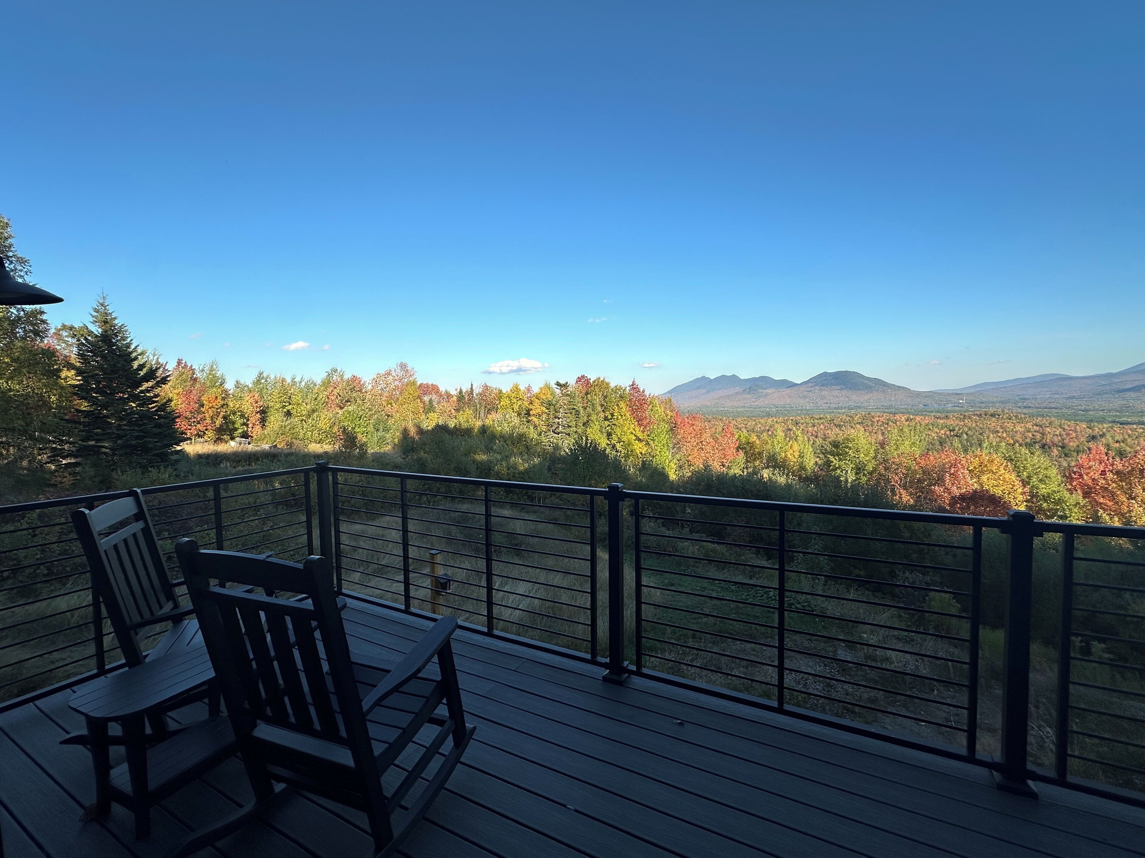 A deck with two wooden chairs and a table, overlooking a forested landscape with autumn foliage and distant mountains.