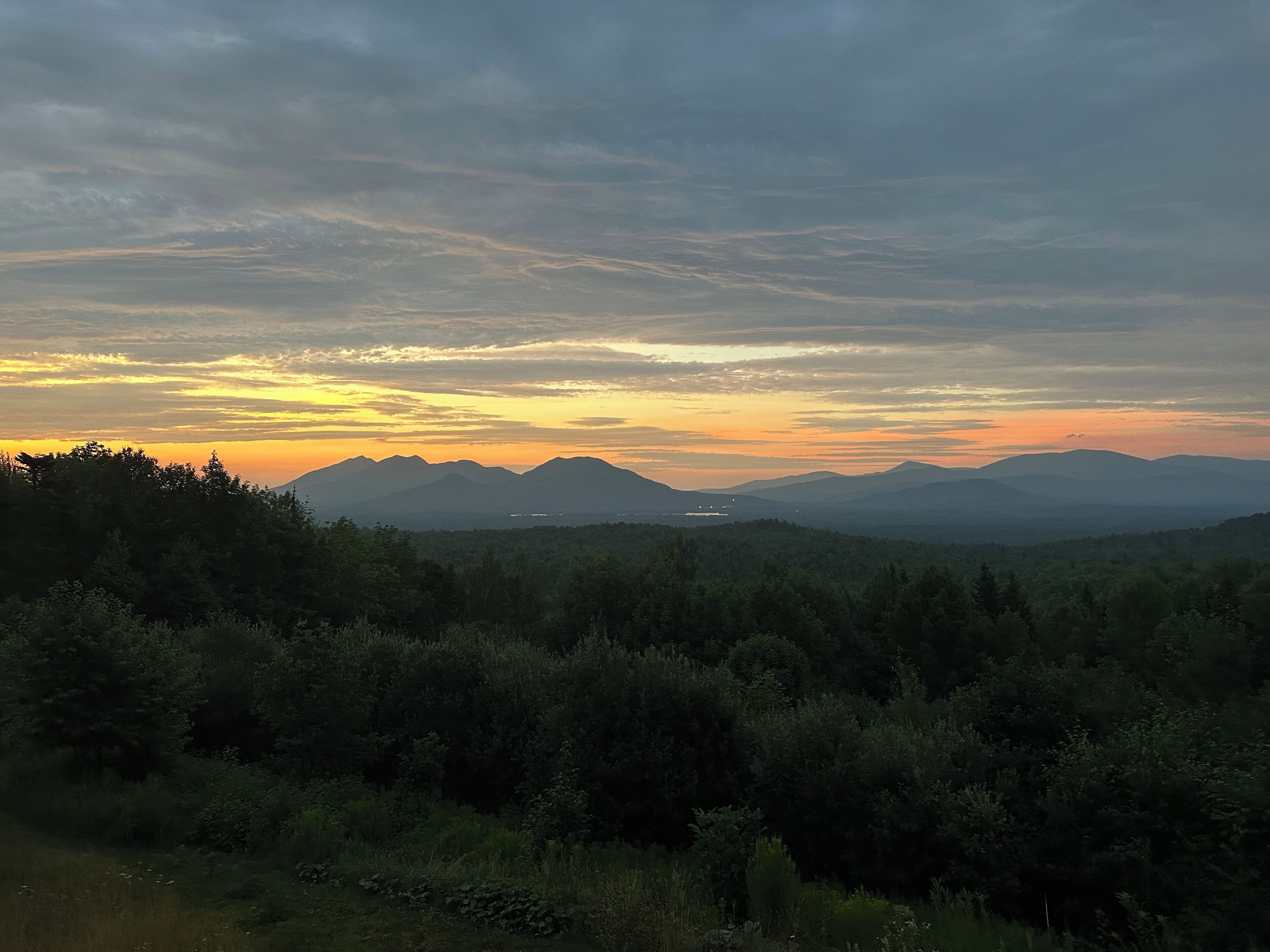 A mountainous landscape with a forested foreground and a sunset sky.