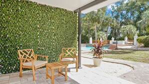 Area esterna con sedute: due sedie di legno e un tavolo, una parete verdeggiante con vegetazione, e vista su una piscina e palme.