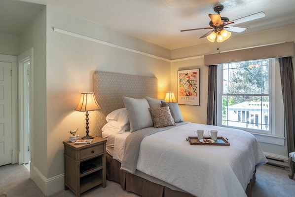 Une chambre bien rangée, avec un lit, des lampes de chevet, un ventilateur de plafond, une fenêtre avec des rideaux et une œuvre d’art encadrée accrochée au mur.
