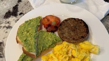 A plate with scrambled eggs, a grilled tomato, a slice of toast with avocado, a sausage patty, and a glass of yogurt with raspberries and mint.