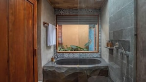 A bathroom with a stone sink, a wooden door, a shower, and a view of greenery through a window. - The Compass Rose Ubud (Bali)