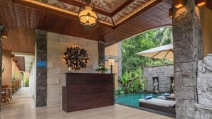 A reception area with a wooden reception desk, a decorative wall piece, and a stone pillar. There is a pool with a stone wall and a statue in the background. - The Compass Rose Ubud (Bali)
