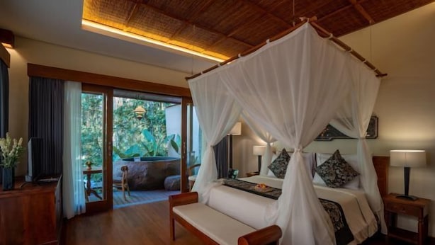 A bedroom with a canopy bed, wooden furniture, a TV, and a view of greenery outside. - The Compass Rose Ubud (Bali)