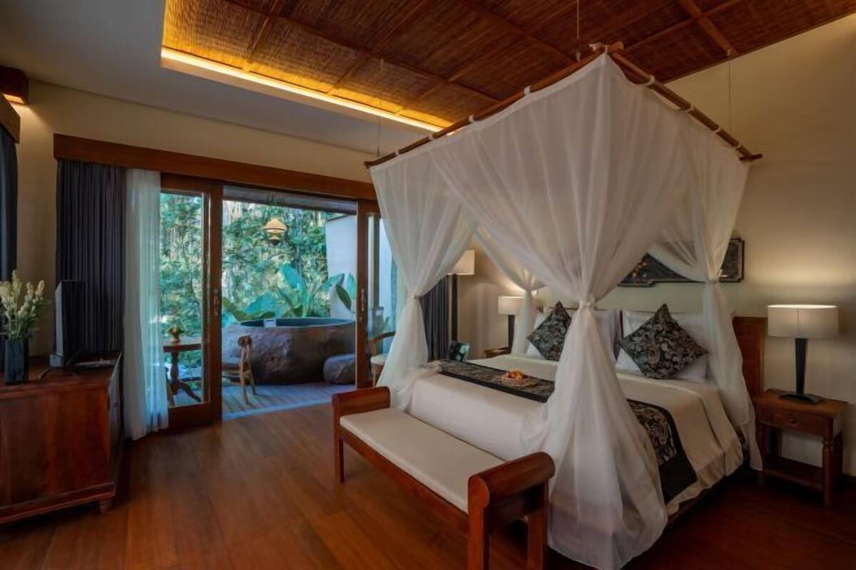 Un dormitorio con cama con dosel, muebles de madera, televisor y vistas a la vegetación exterior.