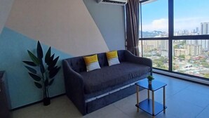 A modern living room with a gray sofa, a small side table, and a large window offering a city view. - Aesthetic 2BR @ Urban Suite (Infinite Pool & Gym) (George Town)