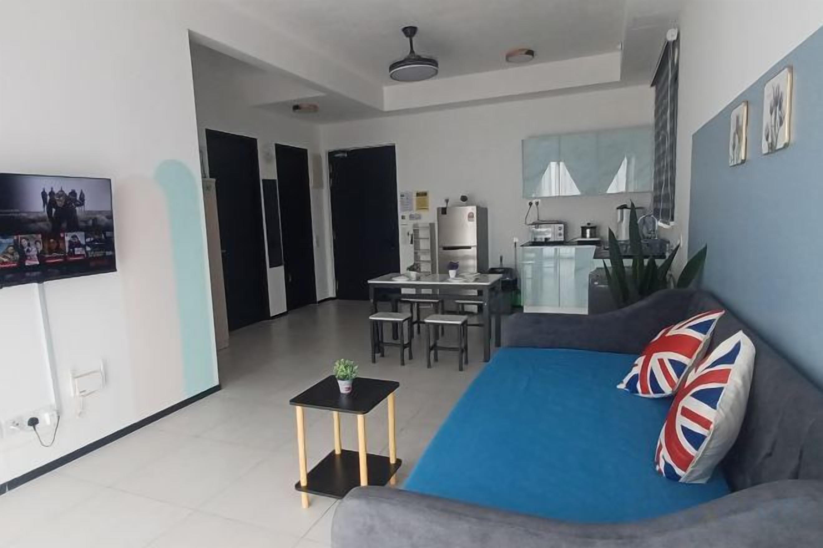 A living room with a sofa, a small table, a TV, and a kitchen area in the background.