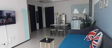 A living room with a sofa, a small table, a TV, and a kitchen area in the background.