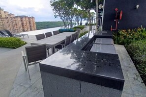 An outdoor seating area with a long table and chairs, a water feature, and a view of a cityscape. - Aesthetic 2BR @ Urban Suite (Infinite Pool & Gym) (George Town)