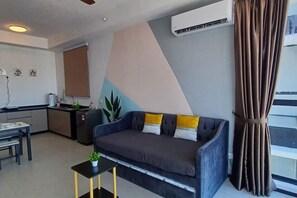 A living room with a sofa, a small table with a plant, a kitchen area, and a large window with curtains. - Aesthetic 2BR @ Urban Suite (Infinite Pool & Gym) (George Town)