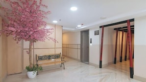 Un hall d’hôtel avec un banc, un arbre en pot, un ascenseur rouge et un sol carrelé.