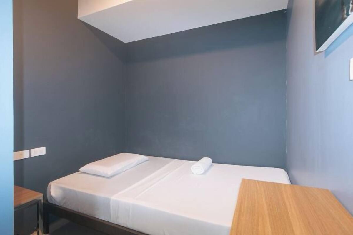 A small, single-bed room with a wooden nightstand and a wall-mounted TV.