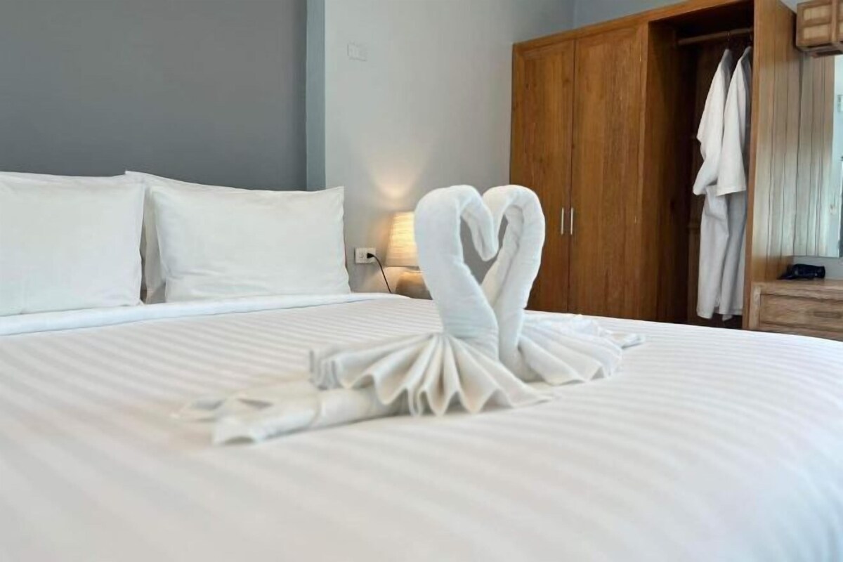A neatly made bed with a towel art in the shape of a heart, a wooden wardrobe, and a mirror on the wall.