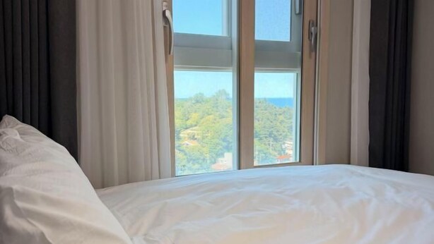 Un lit avec du linge de lit blanc, une fenêtre avec des rideaux et une vue sur la verdure à l’extérieur.