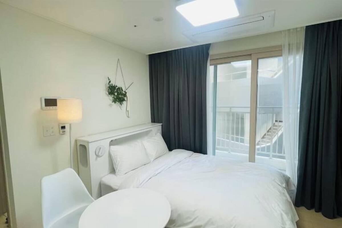 Ein Hotelzimmer mit Bett, Nachttisch, Stuhl, Wandklimaanlage, einem Fenster mit Vorhängen und Blick auf eine Treppe.