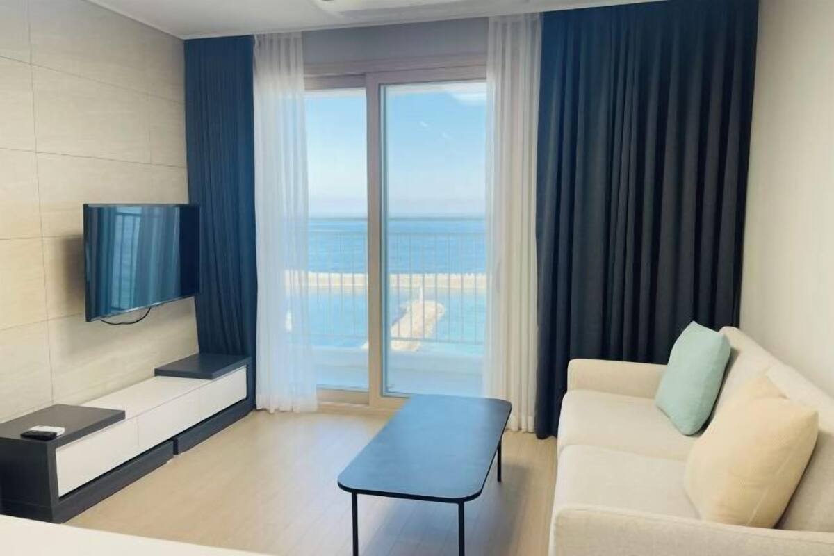 Ein modernes Zimmer mit einer Couch, einem kleinen Tisch, einem Flachbildfernseher und einem Balkon mit Meerblick.