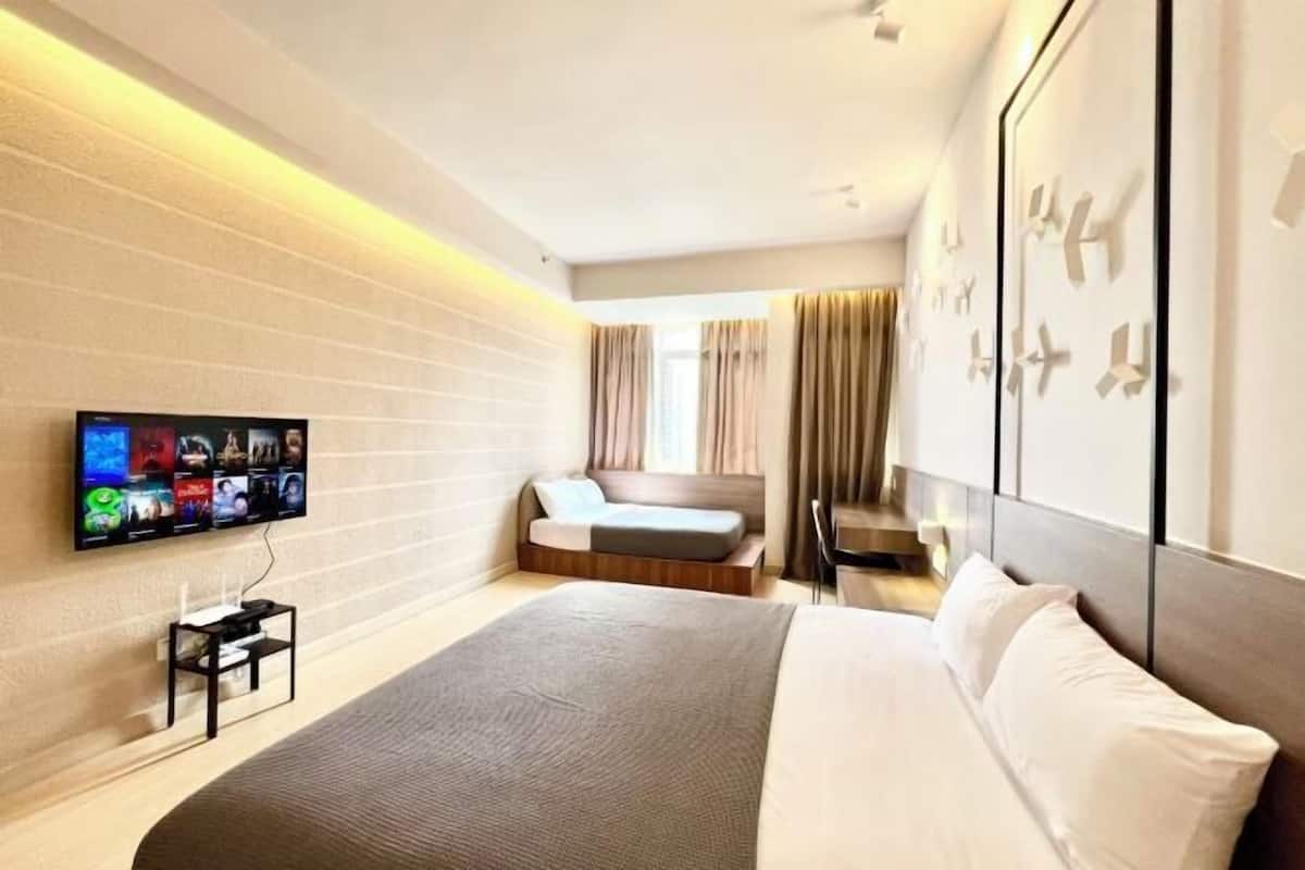A modern hotel room with a large bed, a flat-screen TV mounted on the wall, a small desk, and a seating area with a sofa.