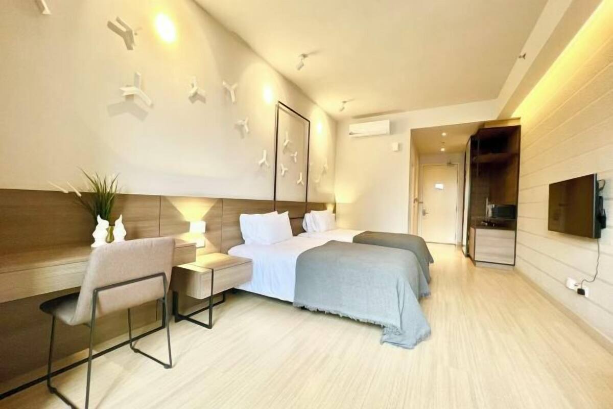 A modern hotel room with a bed, bedside tables, a chair, a TV, and a wall-mounted air conditioning unit.