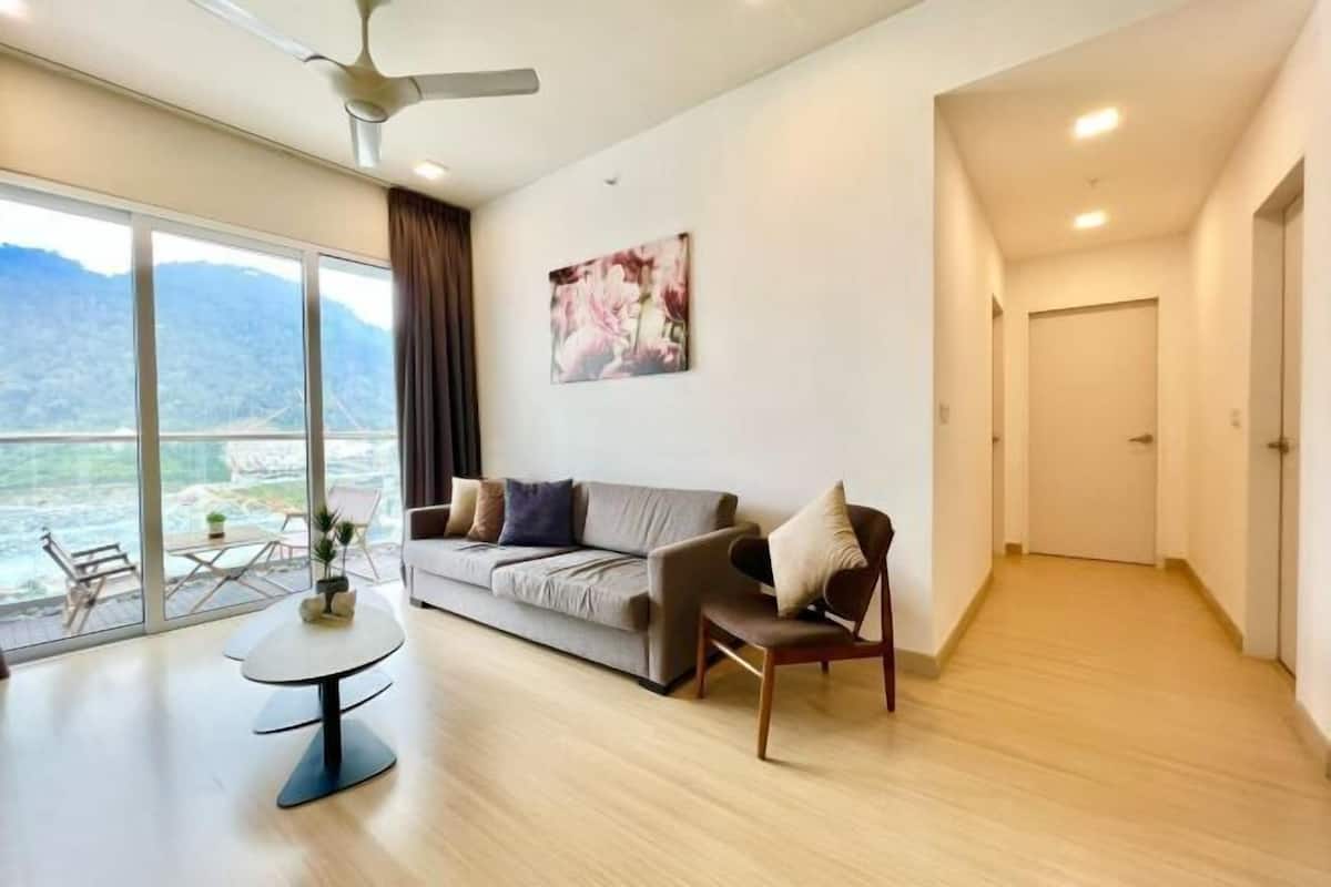 A modern living room with a sofa, a small table, a chair, and a large window offering a view of a body of water and hills.