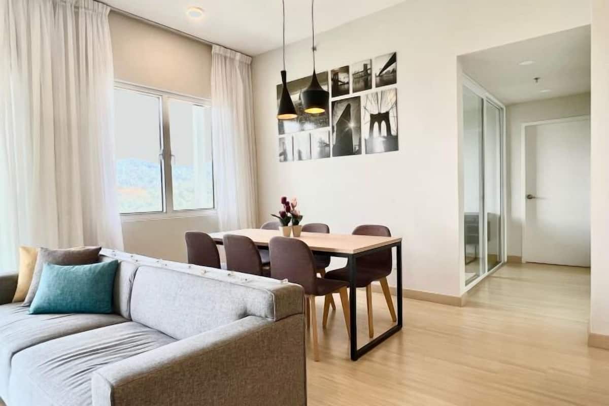 A modern living room with a sofa, dining table, and chairs. There is a large window with curtains, a wall with framed pictures, and a door leading to another room.