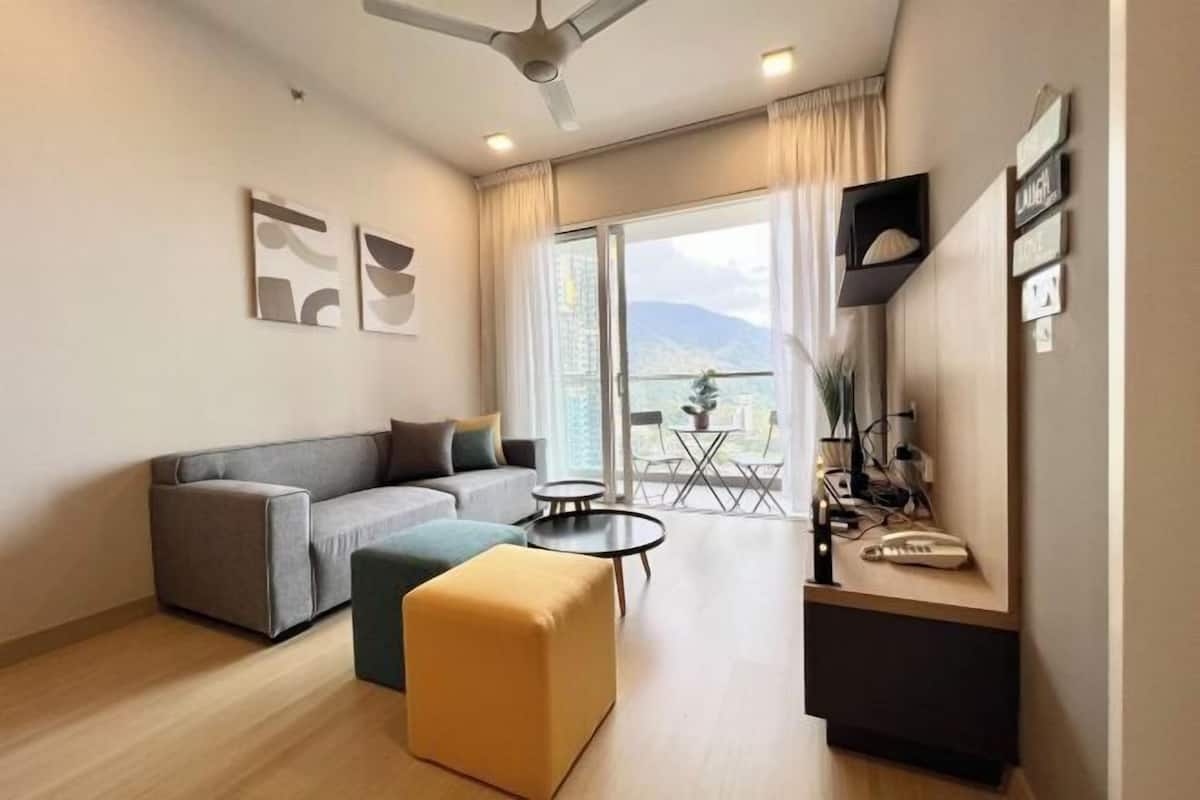 A modern living room with a sofa, ottoman, and a small table. There is a balcony with a view of the mountains.