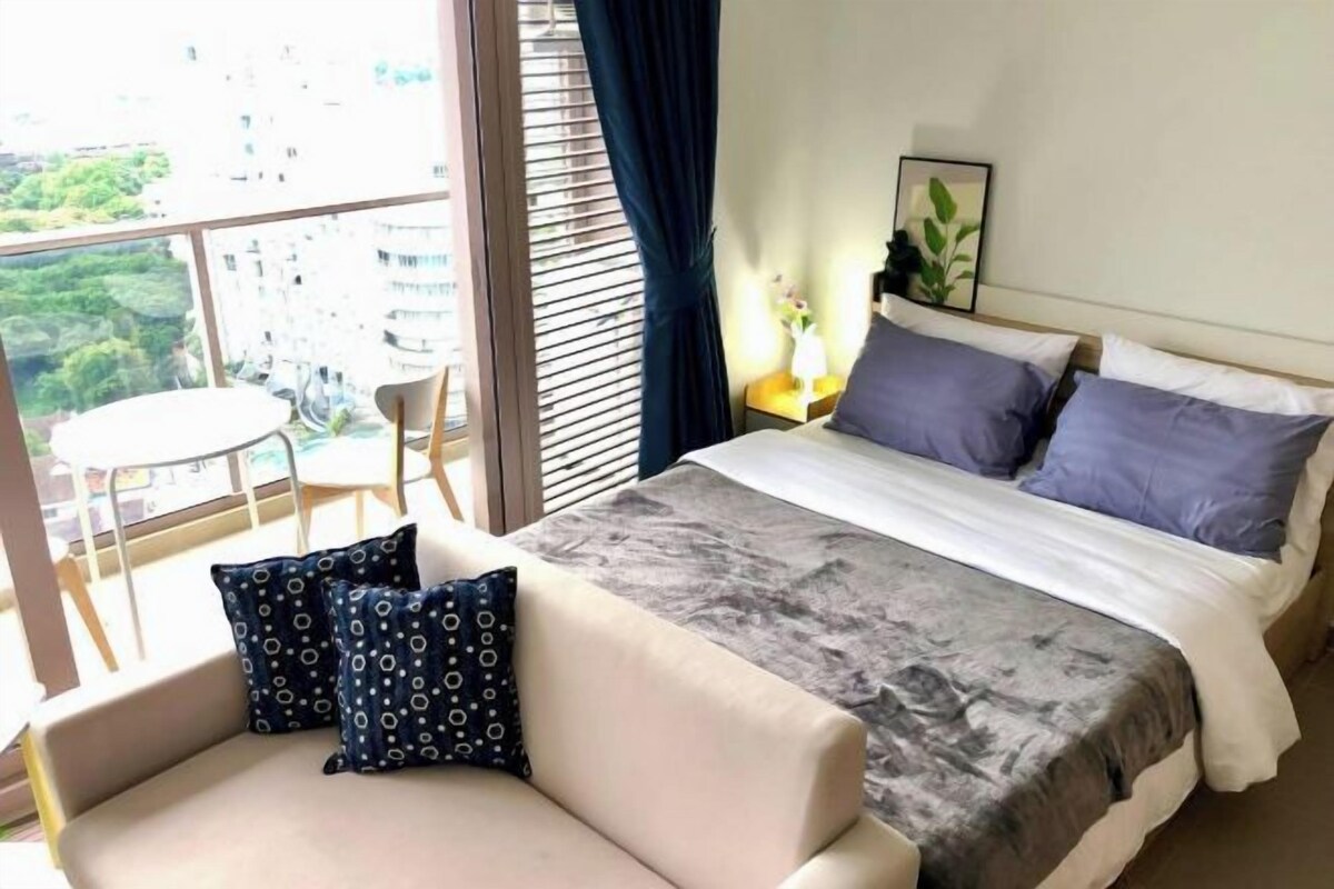 A hotel room with a bed, a sofa, a small table, and a window with a view of buildings and greenery.