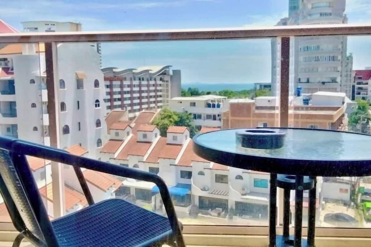 A balcony with a round glass table and a chair, overlooking a cityscape with buildings and a clear sky.