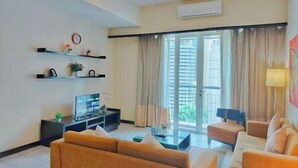 Een moderne woonkamer met een zithoek, een televisie en een salontafel. Er is een groot raam met gordijnen, een airconditioner aan de muur en een klok.