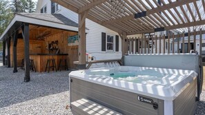 A hot tub under a wooden pergola, a bar area, and a house in the background.