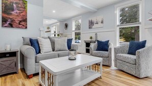 Una sala de estar moderna con un sofá gris, sillones estampados, una mesa de centro blanca y un ventanal con persianas.