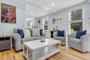 A modern living room with a grey sofa, patterned armchairs, a white coffee table, and a large window with blinds.