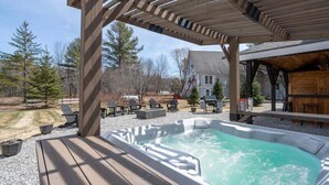 Un jacuzzi bajo una pérgola de madera con mobiliario exterior y una casa al fondo.