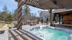 A hot tub under a wooden pergola with outdoor seating and a house in the background.