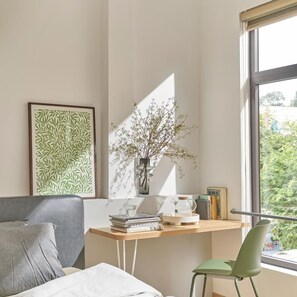 Une chambre moderne avec un lit, un bureau avec des livres et un vase de fleurs, et une fenêtre donnant sur un paysage verdoyant.