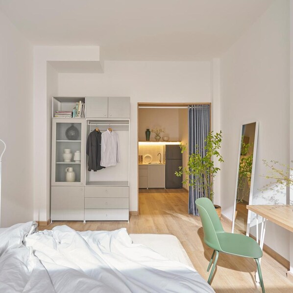 A bedroom with a bed, a green chair, a desk, a wardrobe, and a view into the kitchen.